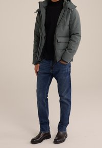 En person står i en ljust studio och har på sig en mörk tröja, en grön puffjacka, blåjeans och svarta stövlar.