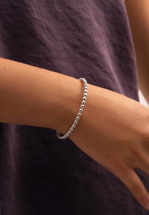 Pulsera de plata con cuentas esféricas de acabado pulido. Lleva en la muñeca, complementada por un fondo de tela texturizada en color púrpura.