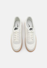 Para białych skórzanych sneakersów Nike z gumowymi podeszwami, białymi sznurówkami i czarnymi logo Nike na języku i wkładkach, widziana z góry.