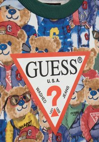 Sweatshirt in cotone con una stampa colorata di un orso in vari outfit, accompagnata da un prominente logo triangolare rosso con la scritta "GUESS U.S.A. WASHED JEANS."