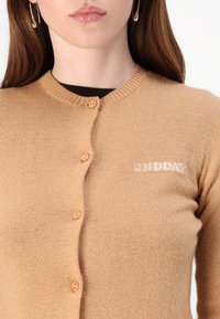 Beige strikket cardigan med rund hals, fem knapper og en liten hvit logo på venstre side. Myk tekstur og figursydd design.