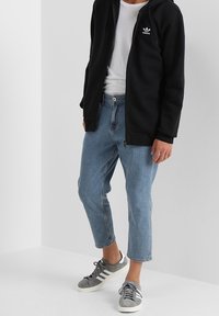 Person som bär avklippta ljusblå jeans, grå Adidas-sneakers, vit t-shirt och svart Adidas zip-up jacka, stående mot en vit vägg.