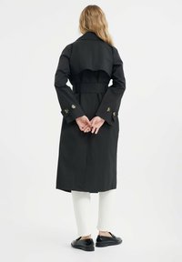 Czarny płaszcz trench z paskiem, detalami w formie peleryny na ramionach, z guzikami na mankietach oraz długości do połowy łydki, w zestawieniu z białymi spodniami i czarnymi butami.