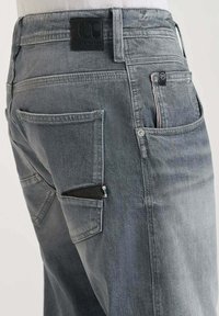 Lichtgrijze denimbroek met een gestructureerde afwerking, meerdere zakken, een zwarte merkpatch en een subtiele rood-wit-blauwe accent op een van de zakken.