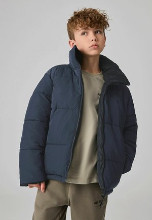 Merinotakki tummansinisellä värillä, korkea kaulus, tikattu muotoilu ja vetoketjukiinnitys. Yllä vaaleanharmaa paita ja beige housut. Pehmeä rakenne.