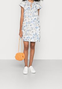 Blumenkleid mit blauen und beigen Mustern, kurzen, flatternden Ärmeln, gerafftem Ausschnitt mit Bindeband, kombiniert mit einer runden orangefarbenen Tasche und weißen Sneakers.