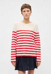 Sark ar garām piedurknēm sarkano un krēmkrāsaino svītru rakstā, ar plašu dekoltē un brīvu piegriezumu, apģērbts ar tumšas džinsa svārku ar krokām.