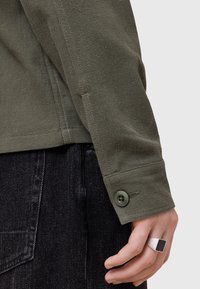 Olivengrøn tekstureret skjorte med knapmanchet. Båret med mørk denim bukser. Synlig ring på hånden. Nærbillede.