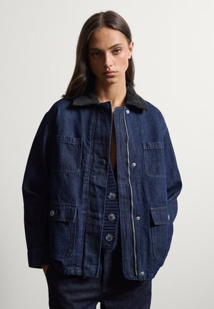 Junge Frau mit langen dunklen Haaren trägt eine dunkelblaue Jeansjacke mit mehreren Taschen über einem geknöpften Stricktop.
