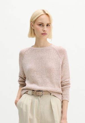 Femme aux cheveux blonds courts portant un pull en tricot rose clair rentré dans un pantalon beige taille haute avec une ceinture, debout les mains dans les poches.