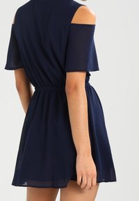 Námořnické modré šaty z lehké tkaniny, s přitaženým pasem, krátkými rukávy a výřezy na ramenou. Sukně má rozšířený tvar.