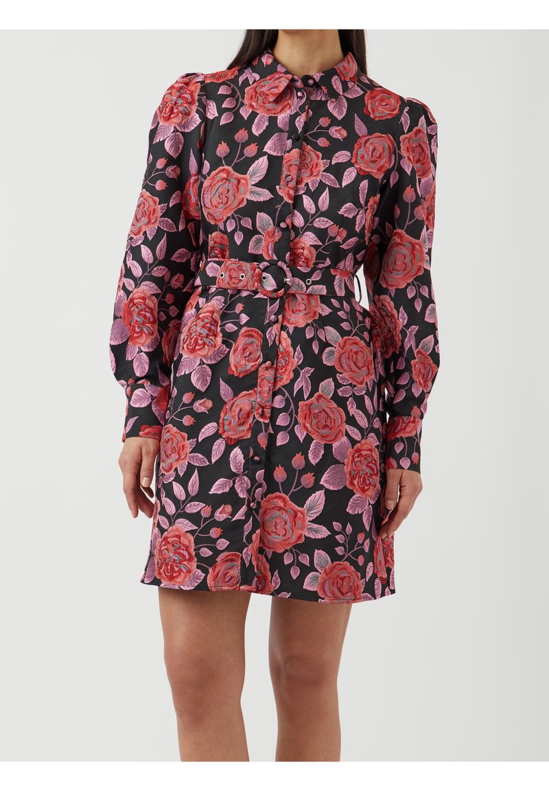 Robe chemise ceinturée noire avec motif floral rouge et rose, manches longues bouffantes et devant boutonné, portée par une femme debout.