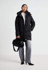 Abrigo acolchado negro con capucha, que presenta un cierre frontal con cremallera, botones a presión y textura acolchada. Combinado con jeans de pierna ancha grises y un bolso negro.