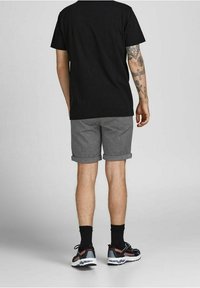 Mężczyzna w czarnej koszulce, szarych podwiniętych szortach, czarnych skarpetkach i trampkach, pokazujący tatuaż na prawej ręce od tyłu.