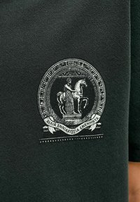 Tamsiai žalia marškinėliai su apvaliu juodai baltu emblema, vaizduojančia arklį ir raitelį, papuošta graikų kalbos tekstu ir dekoratyviniais kraštais.