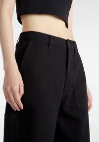 Zwarte wijde broek van gestructureerde stof met voorzakken, riemlussen en een knoopsluiting, weergegeven op taille- en heuphoogte.