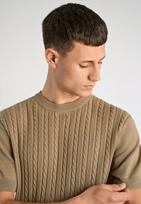 Μπεζ πλεκτό πουλόβερ με υφή σε σχέδιο καλωδίωσης, με ριμπ λαιμόκοψη και κοντά μανίκια. Το ύφασμα φαίνεται μαλακό και ελαστικό.