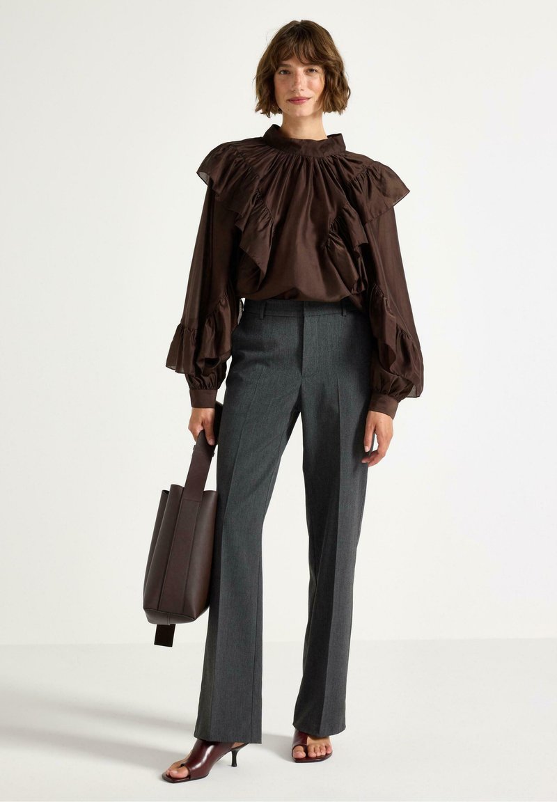 Blusa marrone con volant, colletto alto e maniche lunghe, abbinata a pantaloni grigi sartoriali e una borsa a tracolla marrone scuro. Scarpe con tacco aperte.