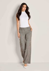 Vrouw met lang, donker haar, gekleed in een mouwloze witte coltrui, grijze wijde broek en witte hakken, staande tegen een lichte achtergrond.