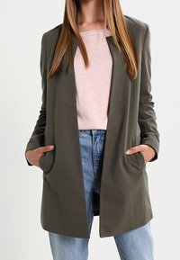 Blazer léger vert olive avec une coupe décontractée, ouverture sur le devant et deux poches latérales, porté par-dessus un haut rose clair et un jean bleu.