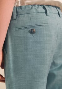 Hellblaue, maßgeschneiderte Hose aus strukturiertem Stoff, mit einer Gesäßtasche und einem kleinen, runden Knopfverschluss.