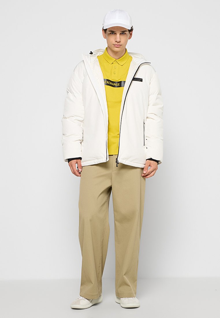 Giacca a vento bianca con cerniere, abbinata a una polo gialla con un logo nero, pantaloni beige e un cappellino bianco. Tessuto liscio, vestibilità rilassata.