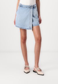 Jupe en jean bleu clair avec une fermeture à boutons sur le devant, des poches latérales et un ourlet épuré, associée à des chaussures à talons ouverts rose pâle.