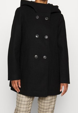 Czarna dwurzędowa marynarka typu pea coat z sześcioma guzikami i kapturem, noszona na beżowych spodniach w kratę, na osobie o średnim odcieniu skóry.