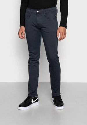 Man draagt strakke donkergrijze broek, zwarte lange mouwen top en zwarte Nike sneakers met witte vleugels en zolen.