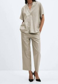 Chemise en soie beige à boutons avec des manches courtes, associée à un pantalon ample assorti et des escarpins noirs à bout pointu, sur fond neutre.