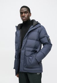 Μπλε τζάκετ πούφφας με κουκούλα, χαρτογραμμένη σχεδίαση, μεγάλες μπροστινές τσέπες και μαύρη εσωτερική επένδυση. Διαθέτει κλείσιμο με φερμουάρ και μανσέτες στους καρπούς.