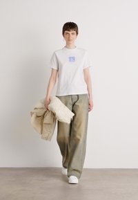 Tricou alb din bumbac cu un logo bleu deschis, asortat cu blugi largi de culoare verde măslinie și o geacă pufos bej. Sneakerși albi completează ținuta.