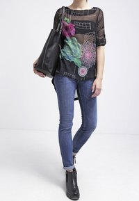 Blouse noire transparente avec des broderies florales colorées et des motifs abstraits, associée à un jean skinny bleu et à un sac fourre-tout noir. Bottines noires.