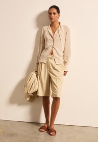 Beige Seidenhemd mit durchgehender Knopfleiste und lockerer Passform, kombiniert mit hellbeigen Shorts. Satinartiger Stoff, braune geflochtene Sandalen und ein passender Blazer.