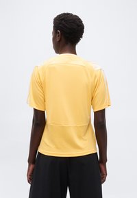 Άτομο με κοντά, σγουρ ά μαλλιά που φορά κίτρινο κοντομάνικο πουκάμισο και μαύρο σορτς, στέκεται πλάτη σε απλό φόντο.