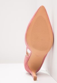 Chaussure à talons hauts en suède rose, à bout pointu, vue depuis la semelle, reposant contre un mur blanc dans un angle.