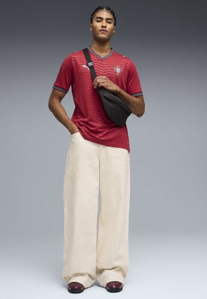 Červený dres s erbem Portugalska, pruhovaná textura. Volné krémové kalhoty, černá kabelka přes rameno a mahagonové boty dokončují outfit.