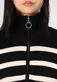 Black and cream striped high-neck sweater with a zipper and circular pull, featuring a ribbed texture and contrasting horizontal stripes.
