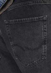 Svarta denimjeans har en bakficka med en enkel, böjd sömdetalj. Texturen är något sliten, vilket ger en subtil djupkänsla.
