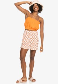 Orange cropped top, ternede shorts i hvid og orange, matchende sandaler og en hat med stribede accenter. Modellen poser med hænderne hævet.
