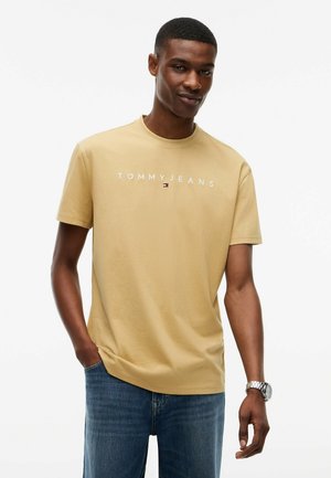 Männliches Model trägt ein beiges Tommy Jeans T-Shirt und blaue Jeans, steht mit einer Hand in der Tasche und einer Uhr am linken Handgelenk.