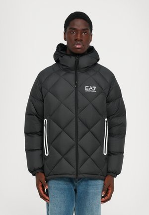 Giovane uomo che indossa una giacca a cappuccio nera trapuntata EA7 Emporio Armani con tasche con zip bordate di bianco e jeans blu, vista frontale.