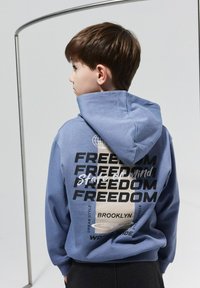 Sinine kapuutsiga, mille seljal on suur print, kus on korduvalt sõna "VABADUS" koos ümmarguse maapalli graafika ja aktsentidega.