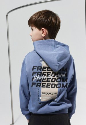 Zils kapucis ar lielu drukātu dizainu, kurā vairākas reizes uz aizmugures ir vārds "BRīvība", ar noapaļota globusa grafiku un akcentiem.