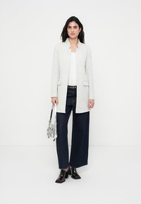 Ανοιχτό γκρι σακάκι με μίνιμαλ σχεδιασμό, χωρίς γιακά, με δύο μπροστινές τσέπες και άνετη εφαρμογή. Συνδυάζεται με σκουρόχρωμο παντελόνι φαρδιάς γραμμής.