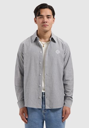 Junger Mann, der ein graues und weißes, vertikal gestreiftes Hemd über einem weißen T-Shirt und blauen Jeans trägt, vor einem schlichten Hintergrund stehend.