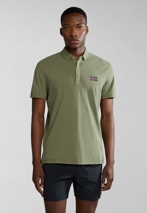 Polo shirt verde oliva con colletto e un piccolo dettaglio ricamato a forma di bandiera sul petto, indossato con pantaloni corti scuri. Texture del tessuto liscia.