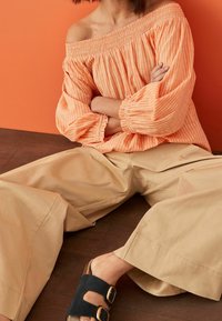Orange and white striped off-shoulder blouse with wide sleeves paired with beige wide-leg trousers and black slide sandals with buckles.