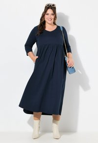 Marineblauwe jurk met lange mouwen, een aansluitende lijfje en een uitlopende rok. Bevat zakken en heeft een V-hals. Gecombineerd met crèmekleurige enkellaarsjes.