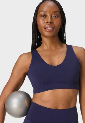 Femme aux cheveux tressés portant un soutien-gorge de sport bleu marine et un legging, tenant un ballon d'exercice argenté contre son côté, fond neutre.